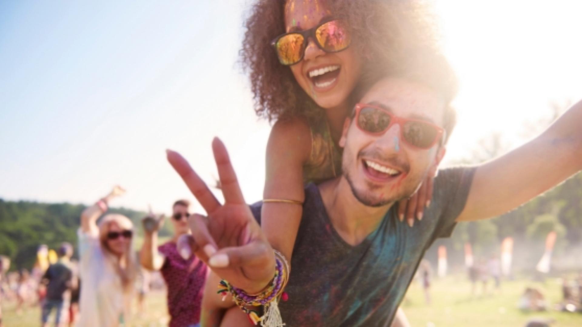 Two people giving each other a piggy-back ride while smiling at the camera in a field with a crowd behind them